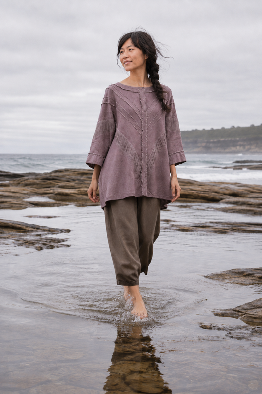 A woman stands barefoot in shallow ocean water on a rocky beach, wearing a loose mauve-purple top and dark brown wide-leg pants under an overcast sky.