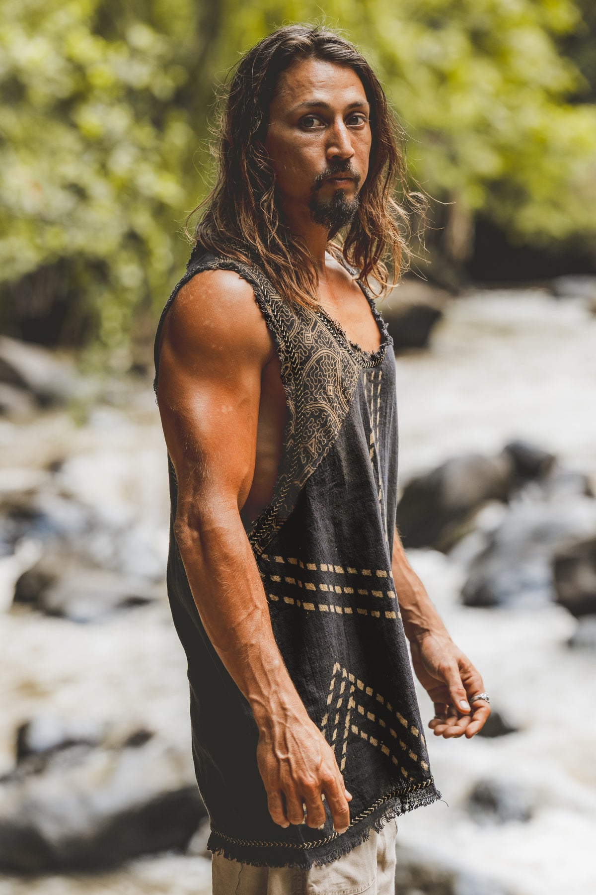Man with long hair and beard standing by a stream wearing a sleeveless garment.