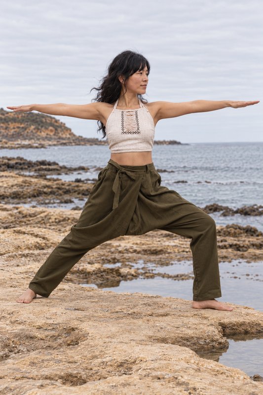 A woman performing a yoga warrior pose on rocky coastal ground with the ocean and cloudy sky in the background. She is wearing an off-white halter crop top and olive green high-waisted loose pants.