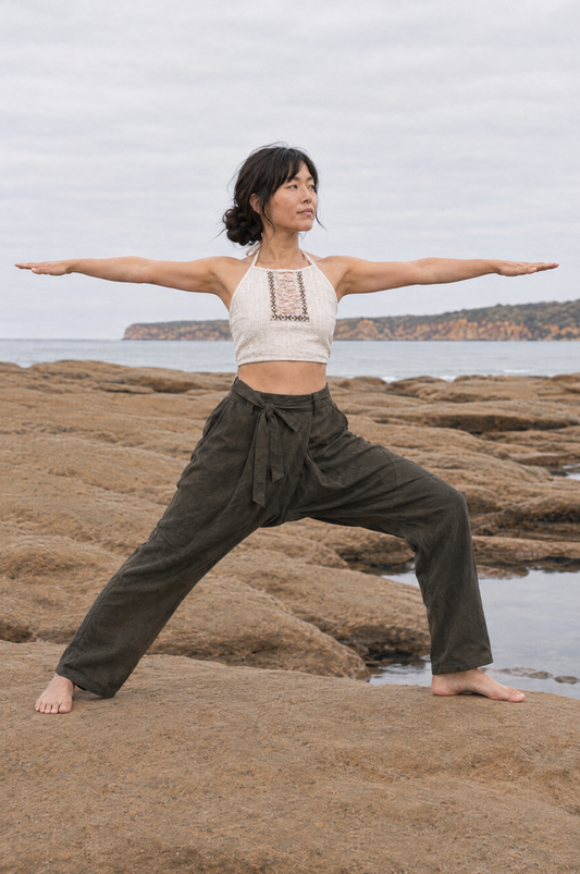A woman performing a yoga warrior pose on rocky coastal ground with the ocean and cloudy sky in the background. She is wearing a cream crochet halter crop top and dark olive green wide-leg pants.