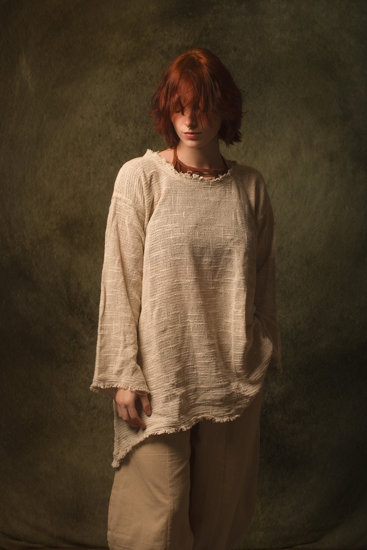A person with short red hair wearing a loose, textured beige top and matching beige pants, standing against a dark olive-green studio backdrop.