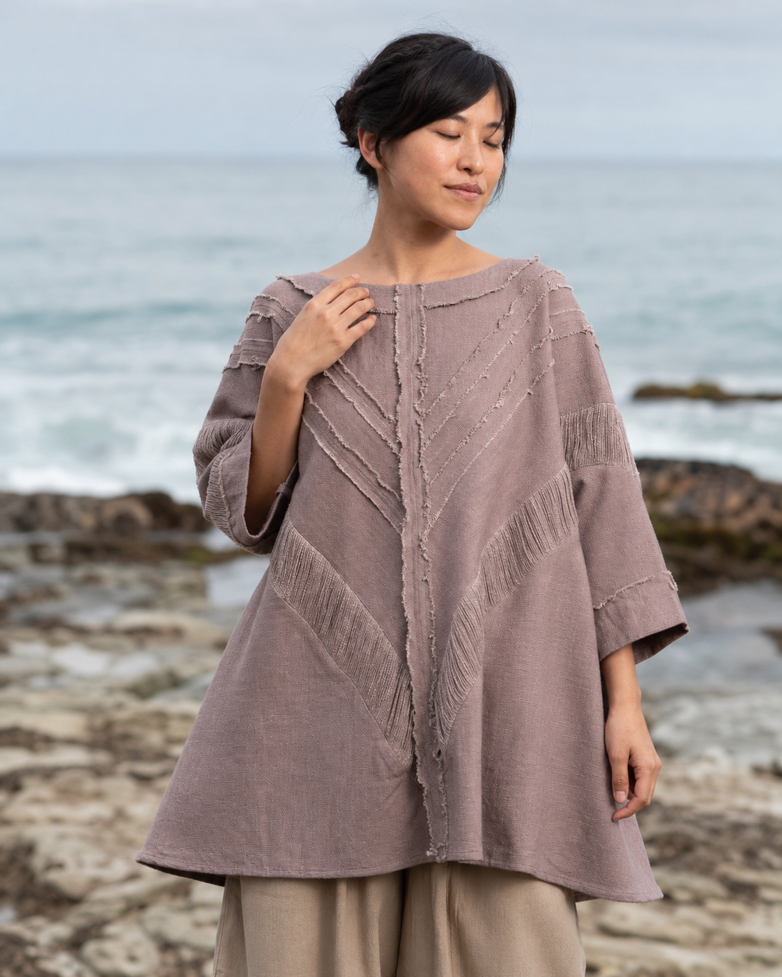 A woman wearing a dusty mauve linen top with textured fringe and chevron details, standing on a rocky beach with the ocean in the background.