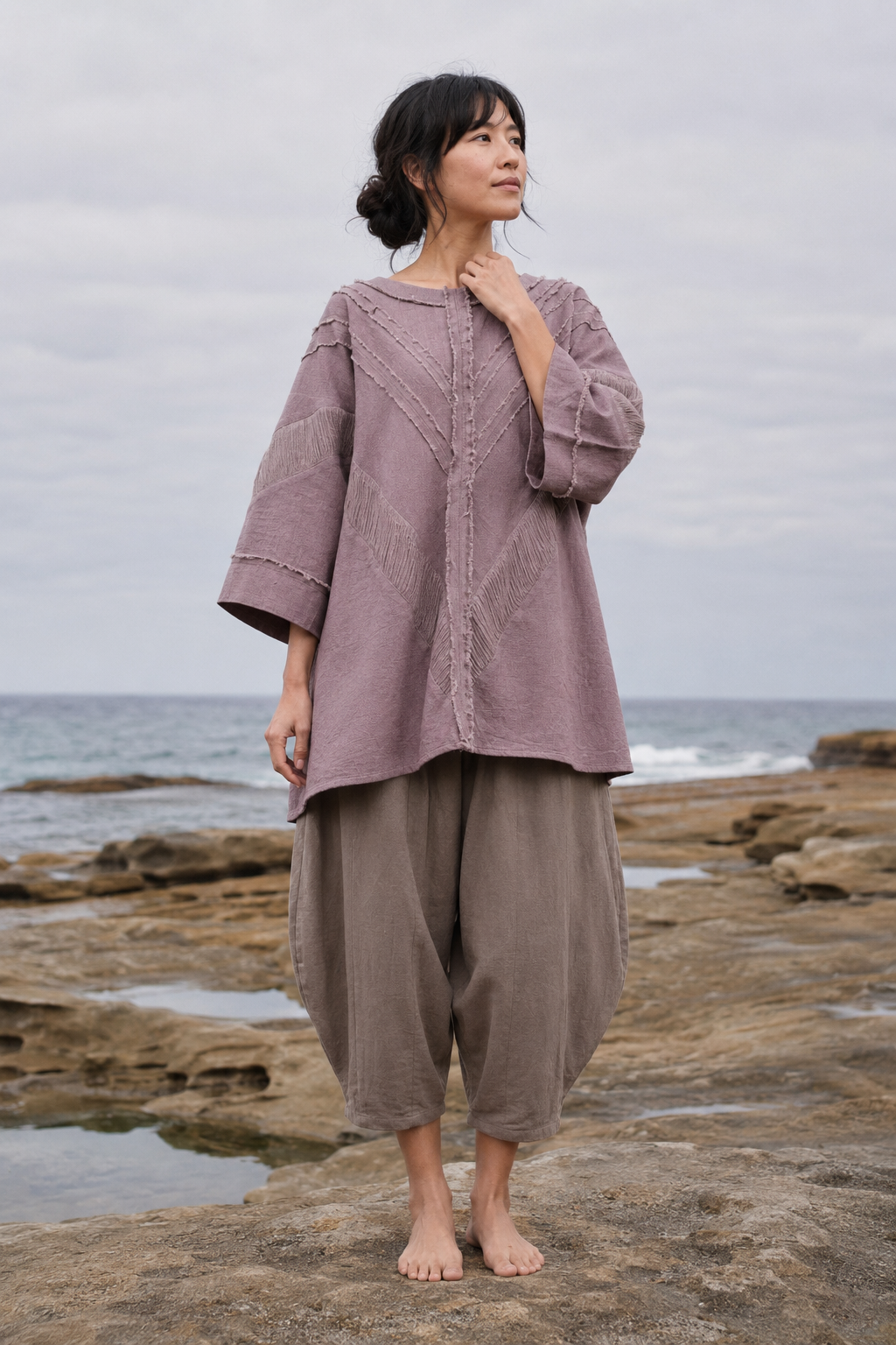 A woman stands barefoot on rocky shoreline with the ocean and cloudy sky behind her, wearing a loose mauve-purple top with three-quarter sleeves and loose taupe-brown wide-leg pants.