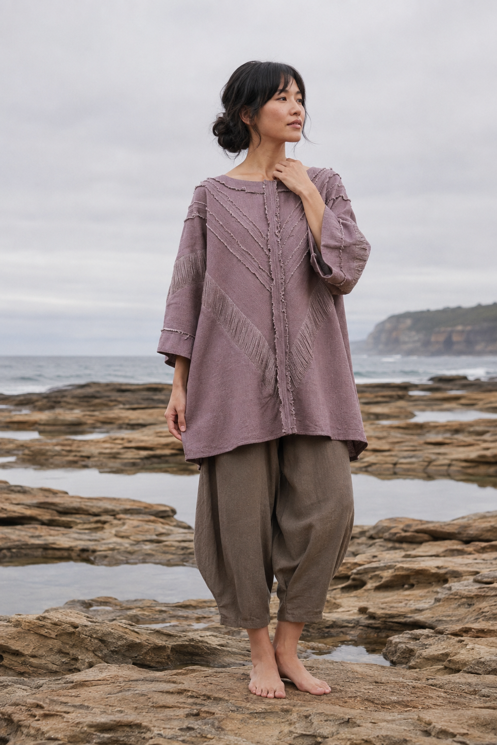 A woman stands barefoot on rocky tide pools at the coast, wearing a loose mauve-lavender top with textured geometric embroidery and three-quarter sleeves, paired with loose taupe pants.