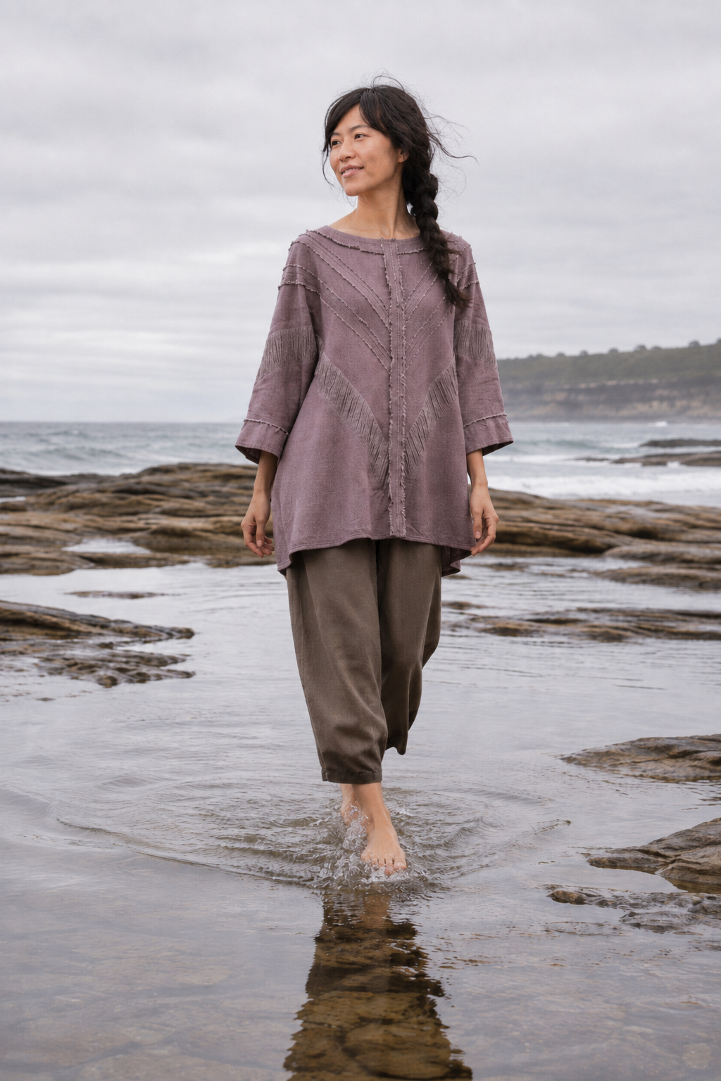 A woman stands barefoot in shallow ocean water on a rocky beach, wearing a loose mauve-purple top and dark brown wide-leg pants under an overcast sky.