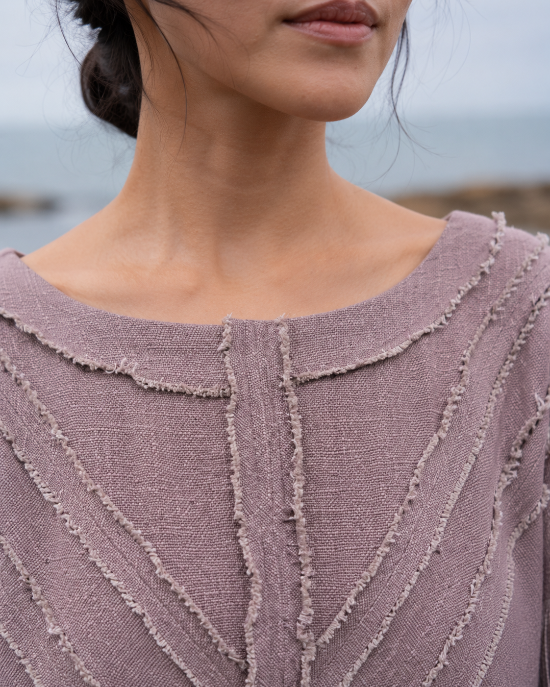 Close-up of a person wearing a textured mauve-pink knit top with frayed trim details, photographed outdoors with a blurred coastal background.