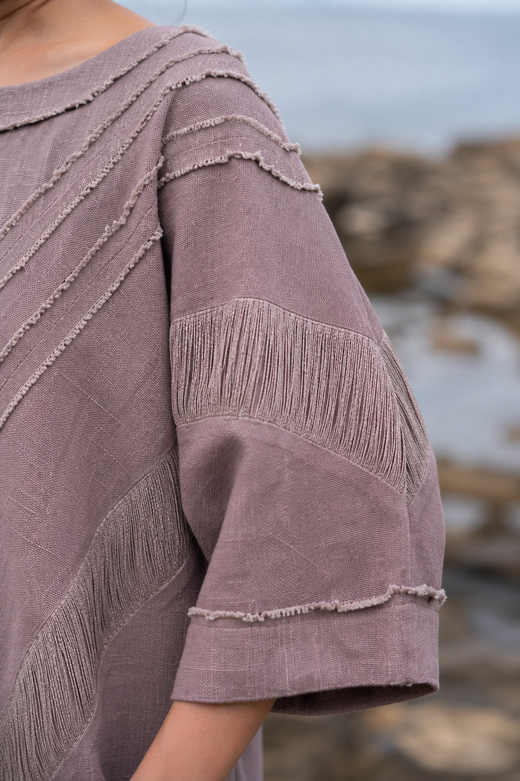 Close-up of a person wearing a dusty mauve linen top with textured fringe and ruffle details on the sleeves, photographed outdoors against a blurred rocky shoreline background.