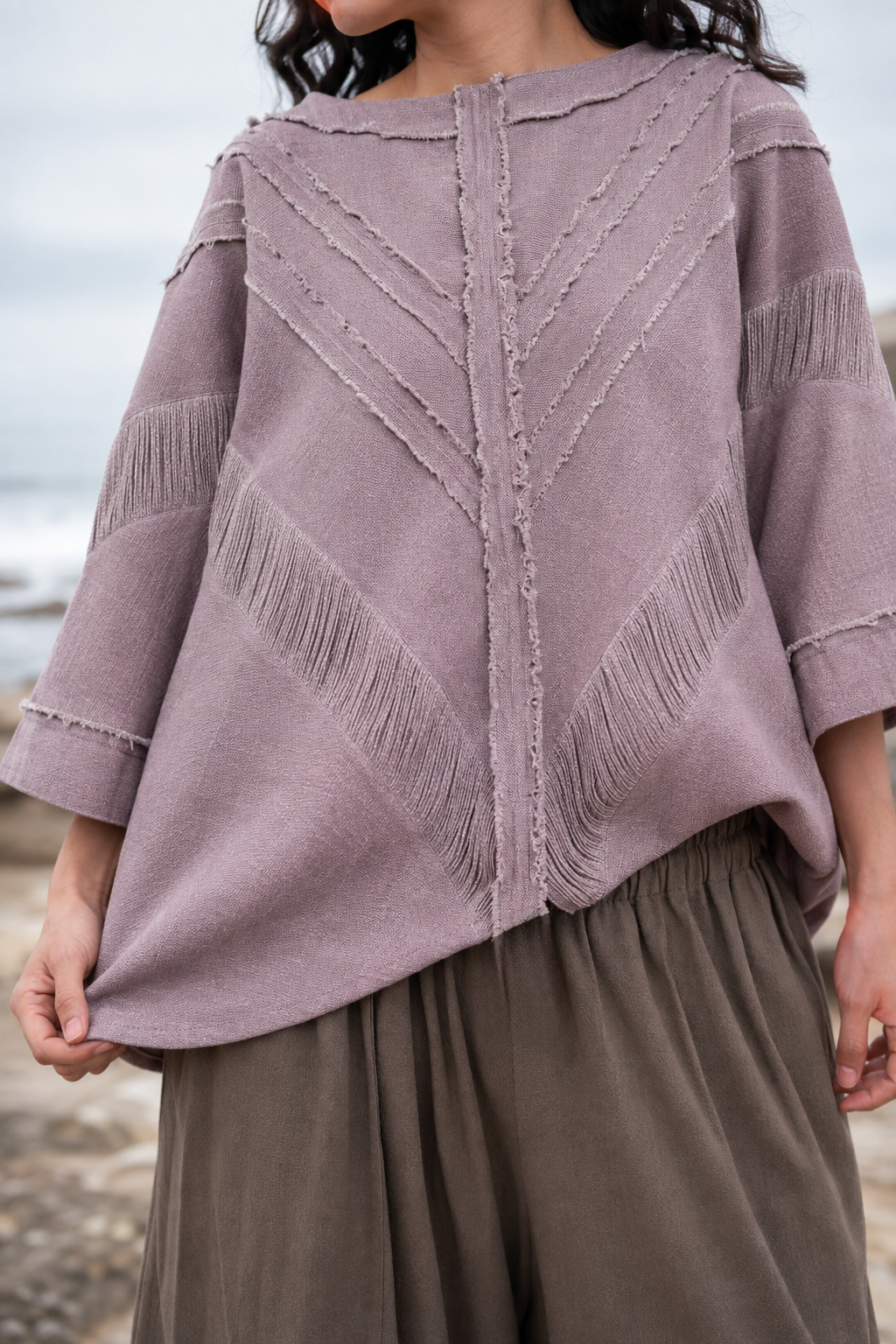 A person wearing a dusty mauve knit sweater with textured fringe and chevron stitching details, paired with brown wide-leg pants, photographed outdoors near the water.