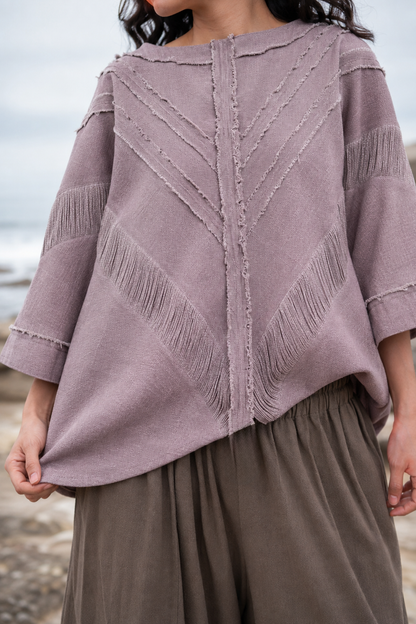 A person wearing a dusty mauve knit sweater with textured fringe and chevron stitching details, paired with brown wide-leg pants, photographed outdoors near the water.