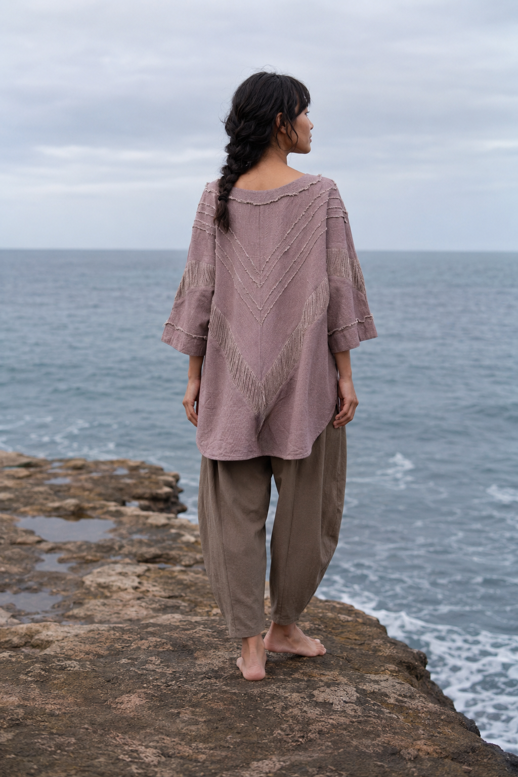 A woman stands on a rocky coast facing the ocean, wearing a loose dusty mauve knit top with three-quarter sleeves and brown wide-leg pants.