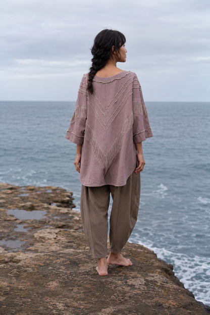 A woman stands on a rocky coast facing the ocean, wearing a loose dusty mauve knit top with three-quarter sleeves and brown wide-leg pants.