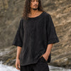 A man with long dark hair stands outdoors against a rocky cliff background, wearing an oversized black textured top with wide three-quarter sleeves and black pants.
