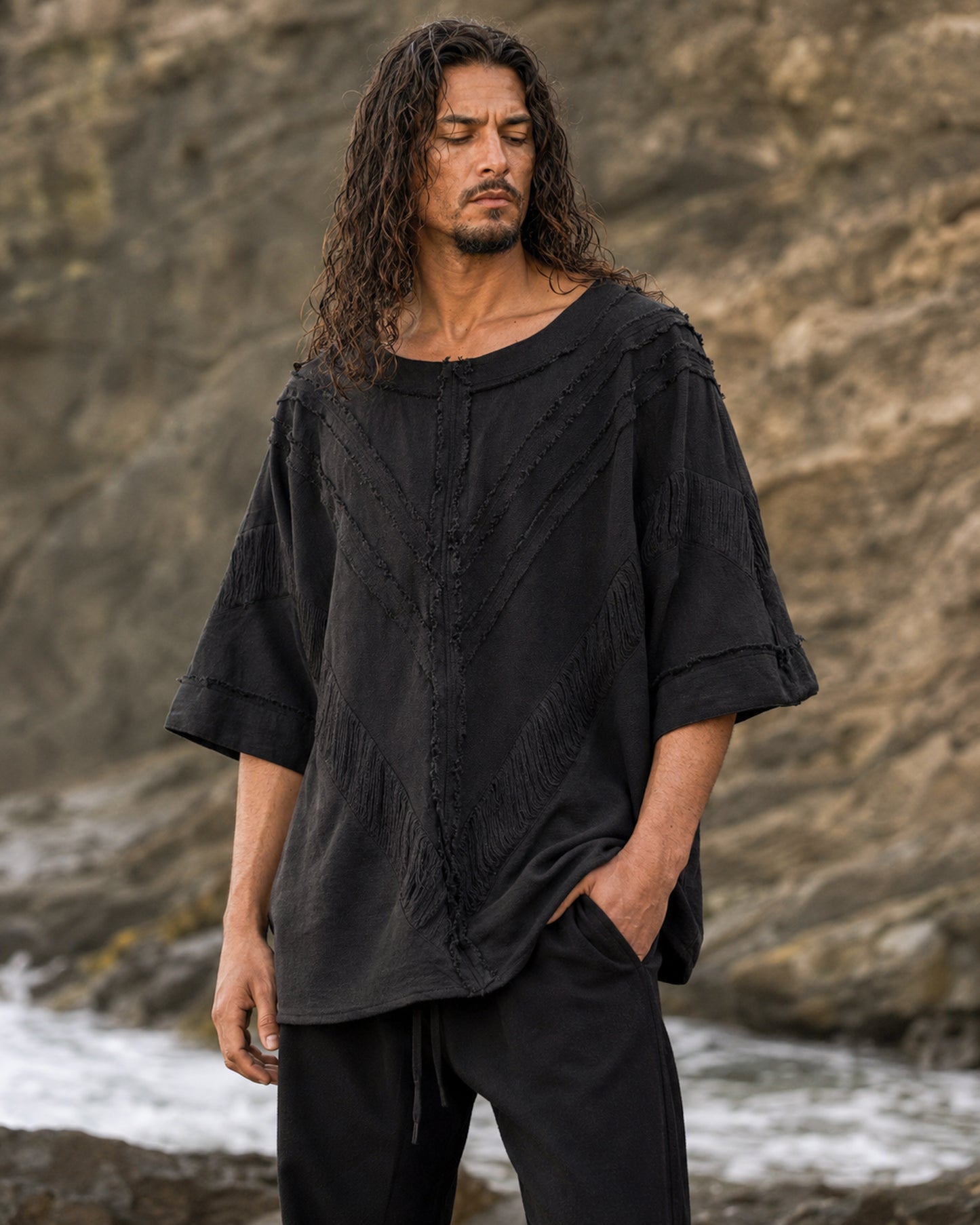 A man with long dark hair stands outdoors against a rocky cliff background, wearing an oversized black textured top with wide three-quarter sleeves and black pants.