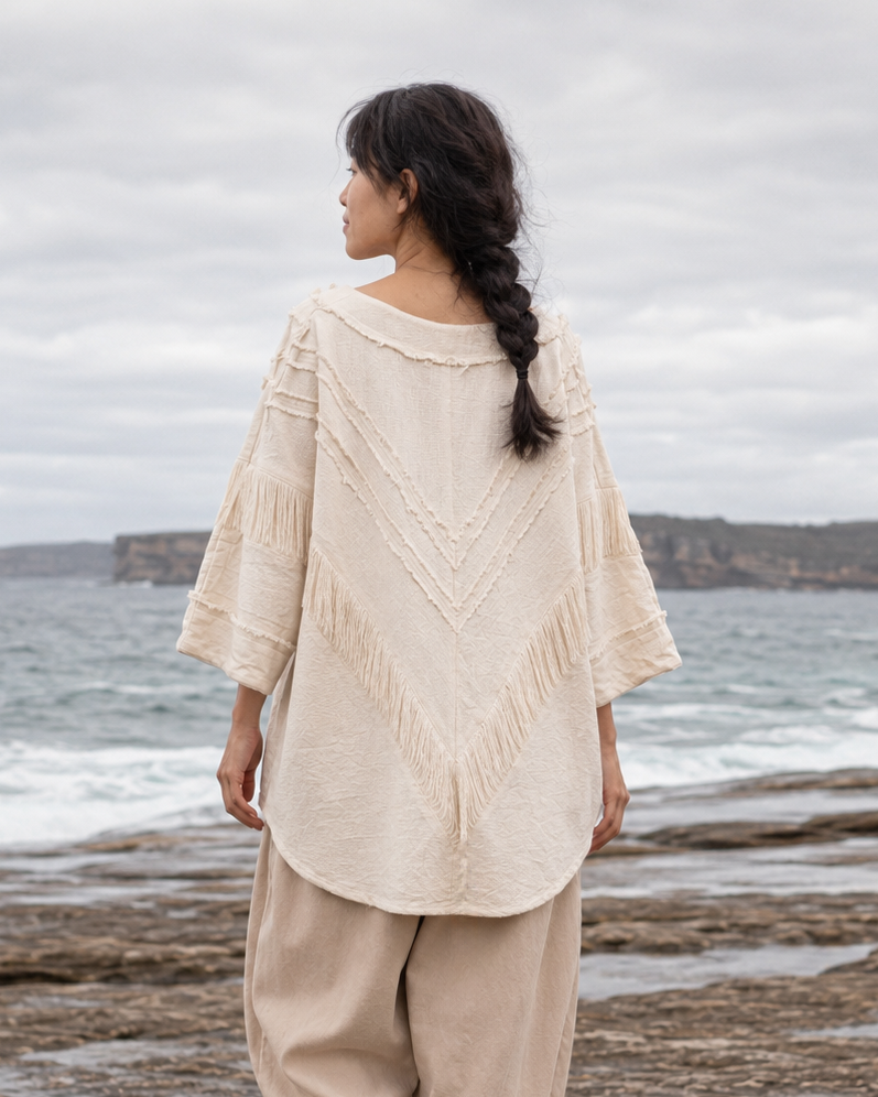 A woman stands barefoot on a rocky beach facing the ocean, wearing a cream-colored, textured bohemian-style top with wide sleeves and matching loose beige pants.