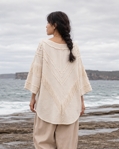 A woman stands barefoot on a rocky beach facing the ocean, wearing a cream-colored, textured bohemian-style top with wide sleeves and matching loose beige pants.