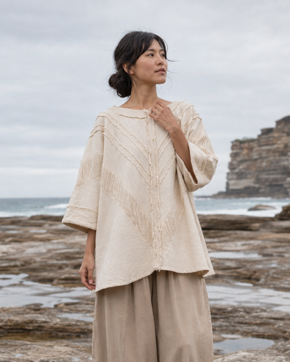 A woman stands barefoot on a rocky ocean shore wearing a loose cream embroidered top and matching beige wide-leg pants, with cliffs and the sea in the background.