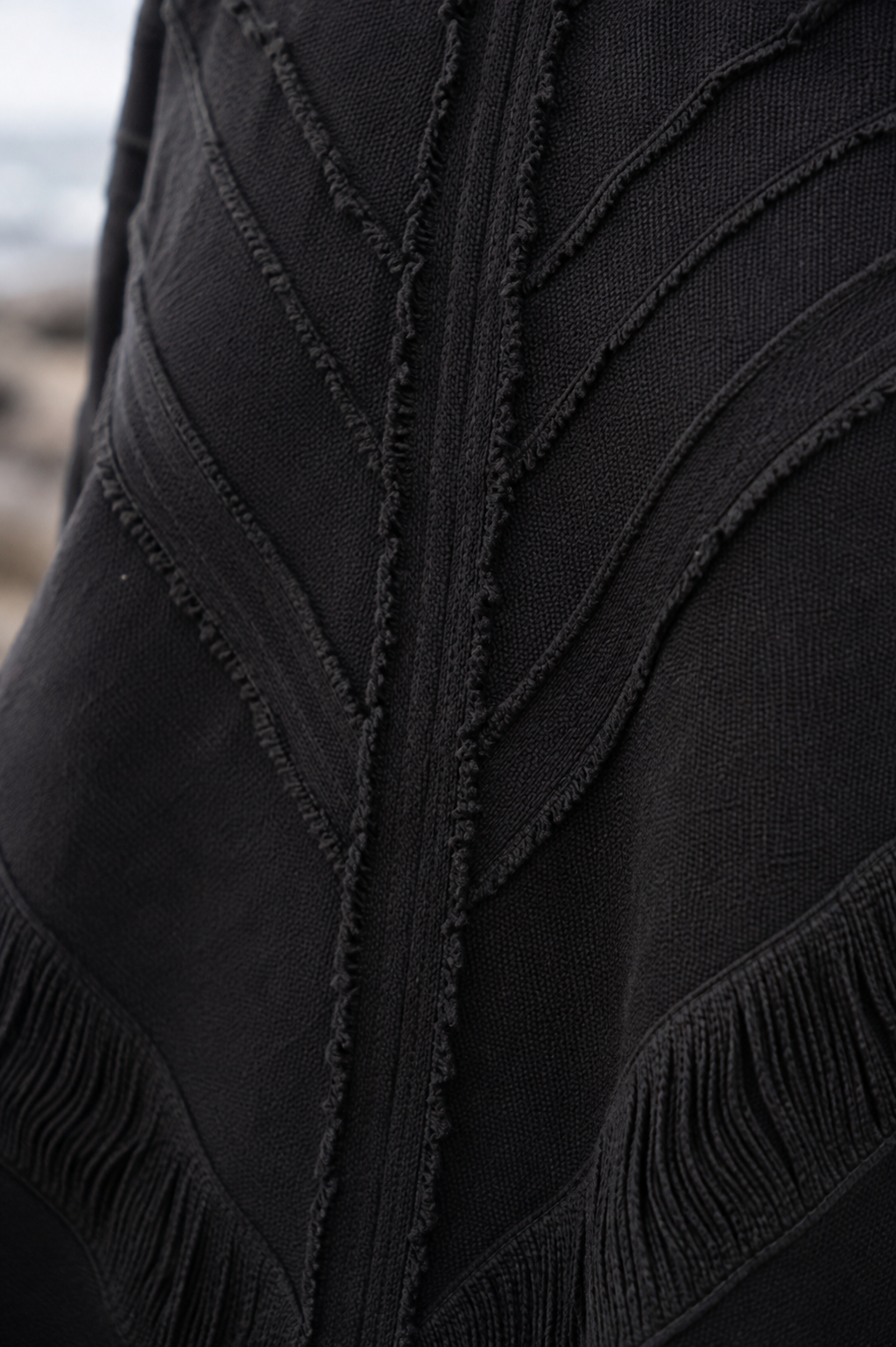 Close-up of a black textured knit garment with leaf-like stitched paneling and fringe trim, photographed outdoors with a blurred natural background.