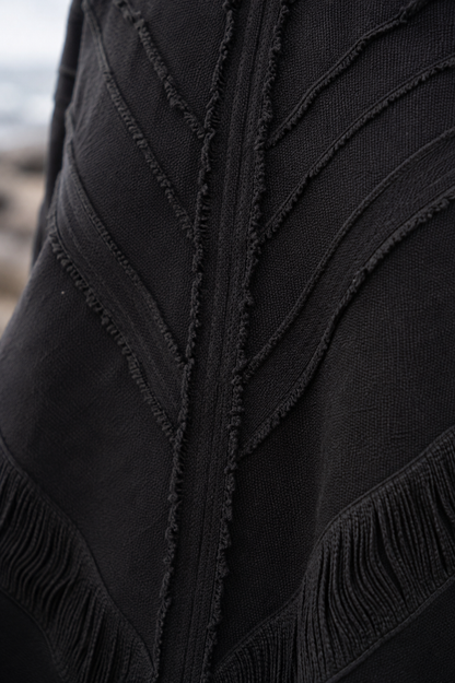 Close-up of a black textured knit garment with leaf-like stitched paneling and fringe trim, photographed outdoors with a blurred natural background.
