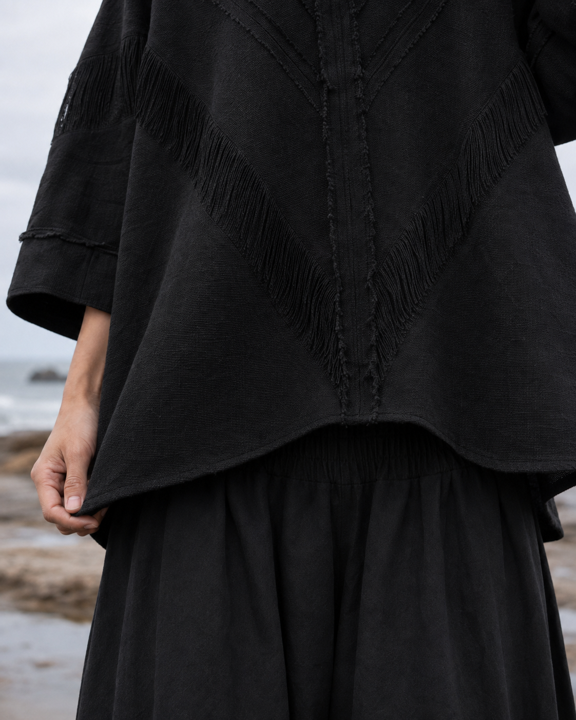 A close-up of a person wearing a black textured top with fringe details and a black skirt, standing outdoors near a beach or rocky shoreline.
