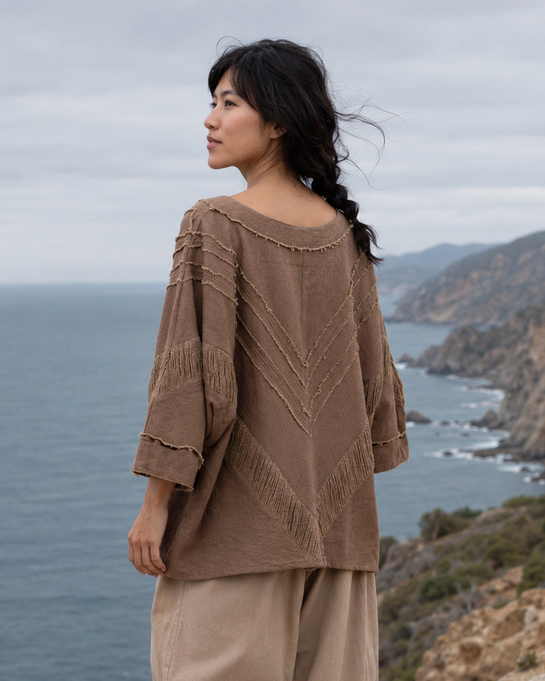 A woman stands barefoot on a rocky coastal cliff, wearing a loose brown bohemian-style top with textured geometric embroidery and beige wide-leg pants, looking out over the ocean.