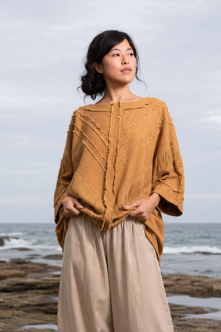 A woman stands barefoot on a rocky shoreline with the ocean and cloudy sky behind her, wearing a loose mustard-yellow textured top and light beige wide-leg pants.