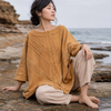 A woman sits barefoot on rocky coastal ground with the ocean and sky behind her. She is wearing a loose mustard-yellow knit top and light beige pants.