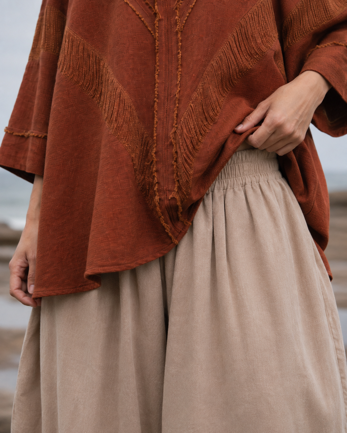 A close-up of a person wearing a rust-orange textured top with fringe trim and beige wide-leg pants, photographed outdoors against a soft blurred background.