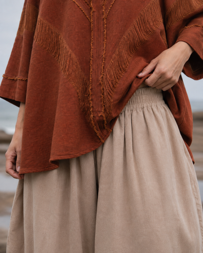 A close-up of a person wearing a rust-orange textured top with fringe trim and beige wide-leg pants, photographed outdoors against a soft blurred background.