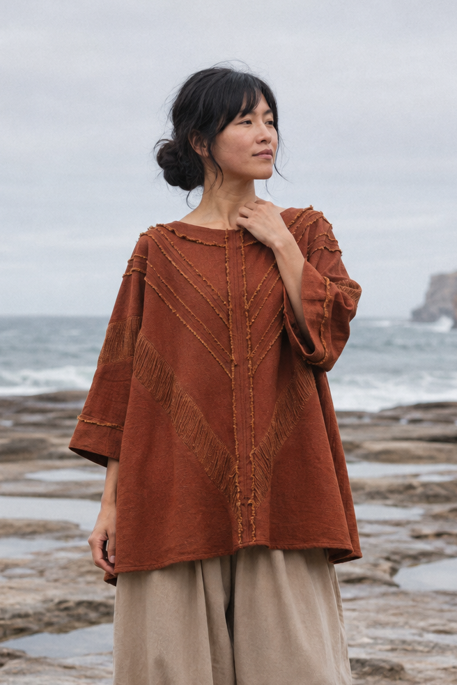 A woman stands barefoot on a rocky ocean shoreline wearing a loose rust-orange embroidered top and beige harem-style pants, with waves and cliffs in the background.
