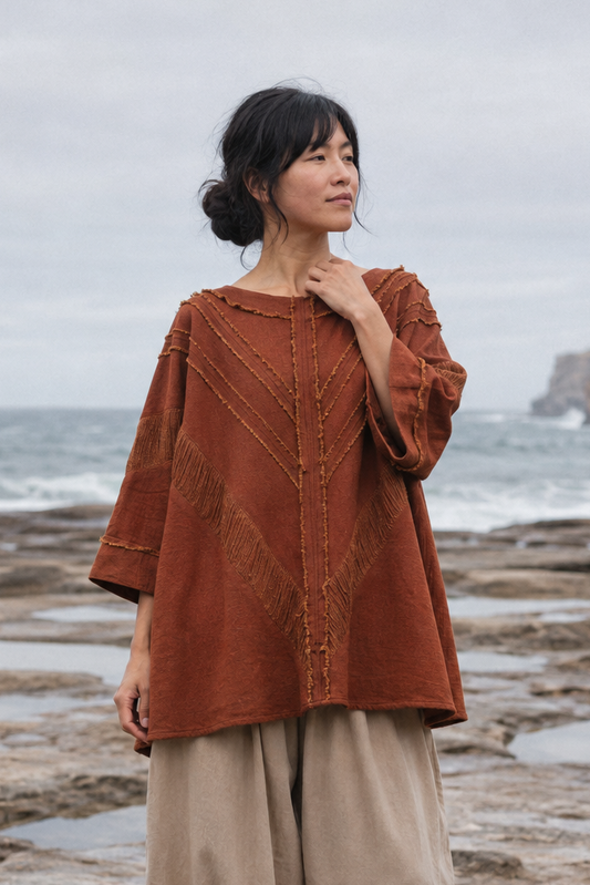 A woman stands barefoot on a rocky ocean shoreline wearing a loose rust-orange embroidered top and beige harem-style pants, with waves and cliffs in the background.