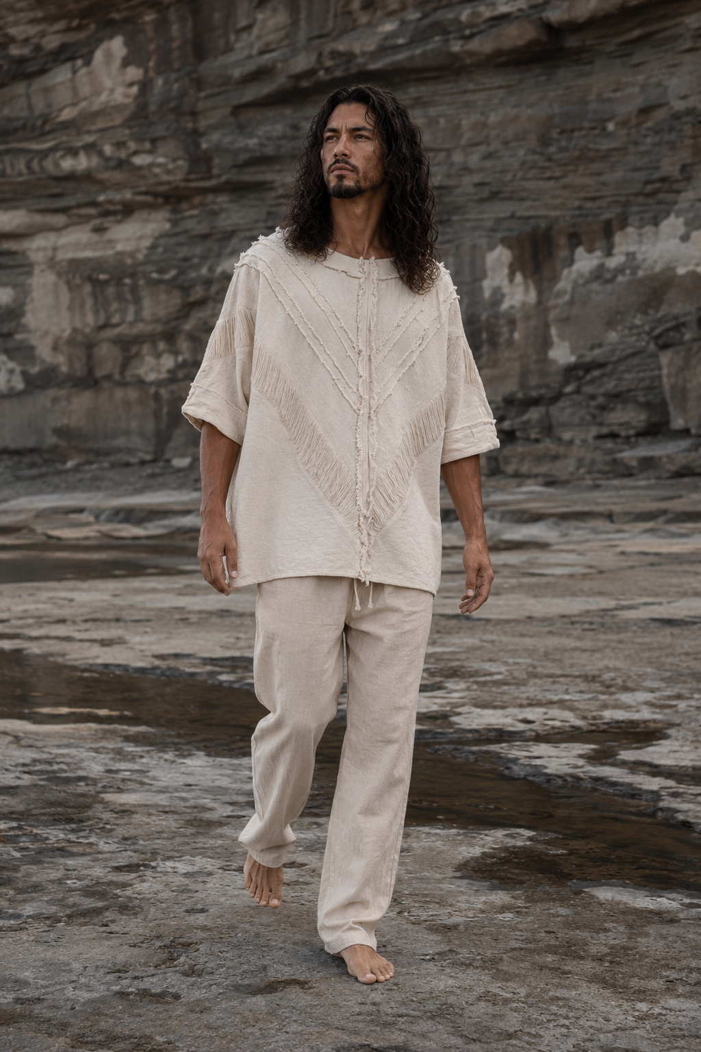 A man with long dark hair walks barefoot on a rocky, outdoor terrain wearing a cream-colored, textured short-sleeve top and matching pants.