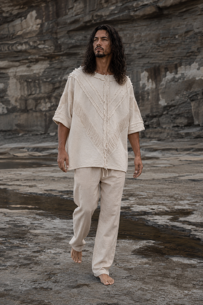 A man with long dark hair walks barefoot on a rocky, outdoor terrain wearing a cream-colored, textured short-sleeve top and matching pants.