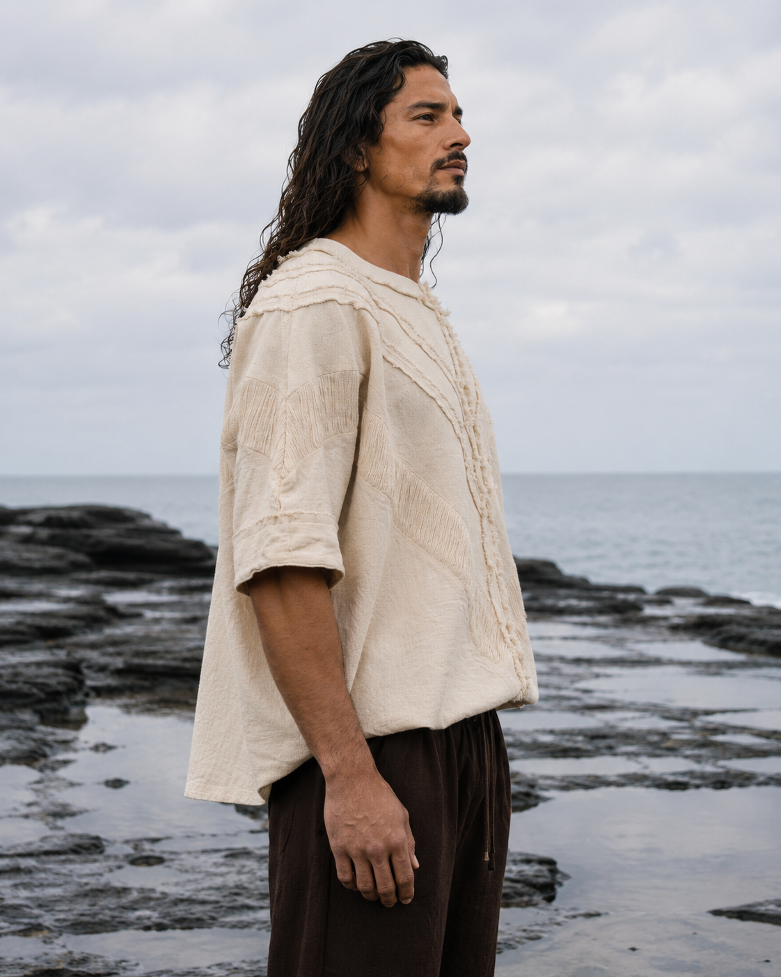 A man with long dark hair stands in profile on a rocky shoreline under an overcast sky, wearing a loose off-white textured short-sleeve top and dark brown pants.