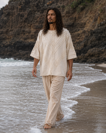 A man with long dark hair walks barefoot along a sandy beach with ocean waves and rocky cliffs in the background, wearing a loose, short-sleeve cream linen tunic and matching linen pants.