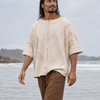 A man walking through shallow ocean water wearing a loose cream-colored short-sleeve top with a textured front and brown pants.