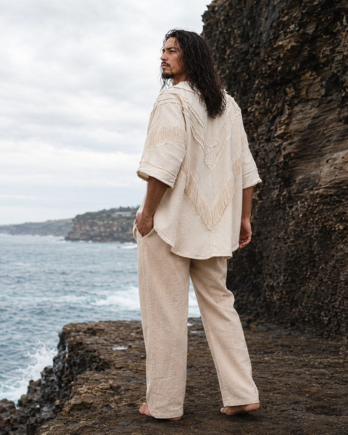 A man with long dark hair stands on a rocky coastal cliff wearing a loose beige linen two-piece outfit with a textured embroidered back design, looking out over the ocean under an overcast sky.