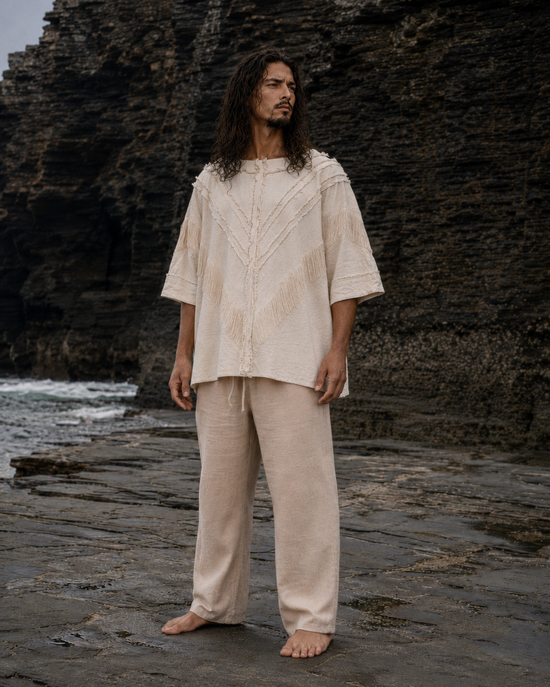 A man with long dark hair stands barefoot on a rocky shoreline wearing a loose, cream-colored two-piece outfit with a textured, hand-dyed pattern. The background features dark, rugged cliffs and wet stone pavement.
