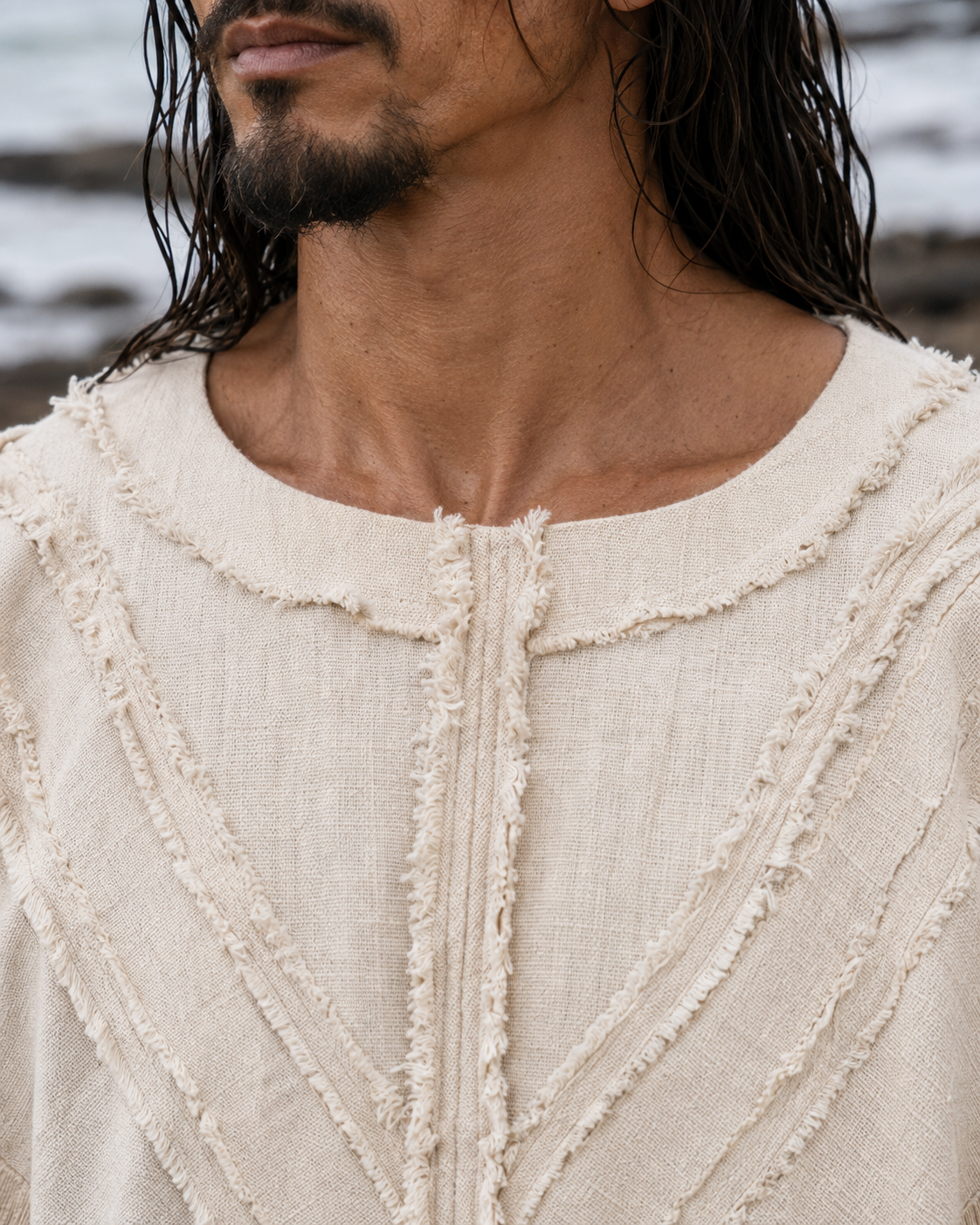 Close-up of a man wearing a cream-colored textured linen top with frayed trim details, set against an outdoor beach background.
