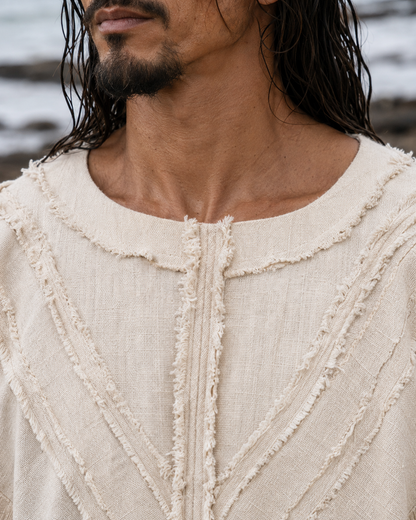 Close-up of a man wearing a cream-colored textured linen top with frayed trim details, set against an outdoor beach background.