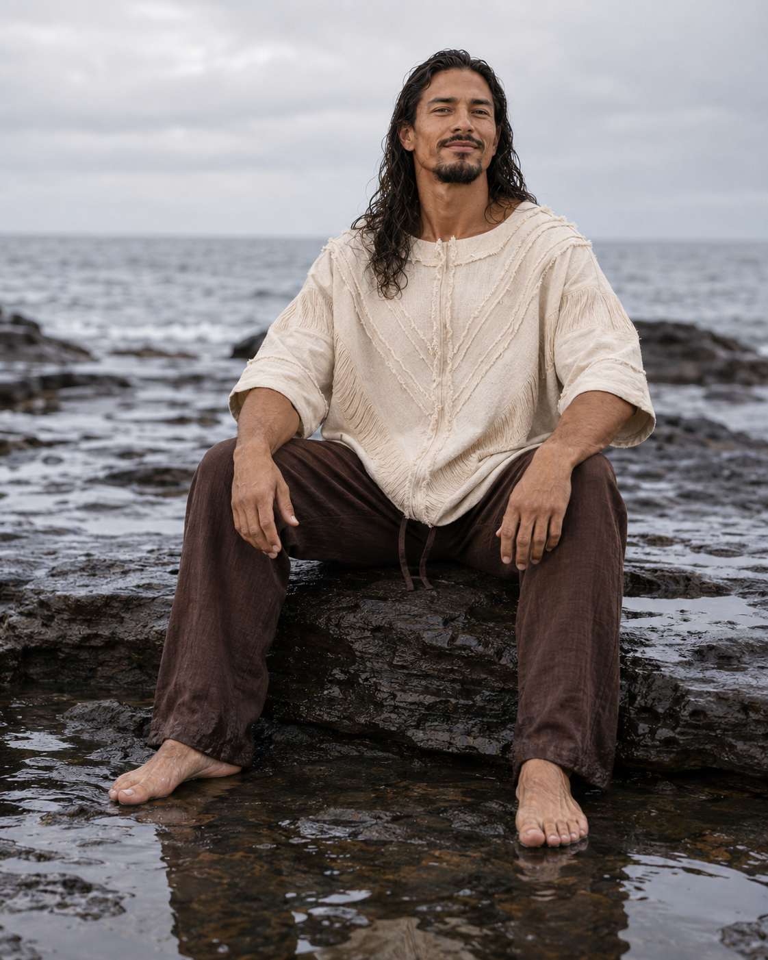 A man with long dark hair sits barefoot on wet rocks by the ocean, wearing a loose cream knit top and dark brown pants.