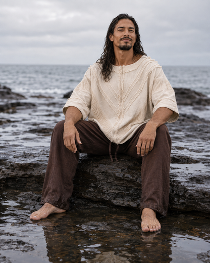 A man with long dark hair sits barefoot on wet rocks by the ocean, wearing a loose cream knit top and dark brown pants.
