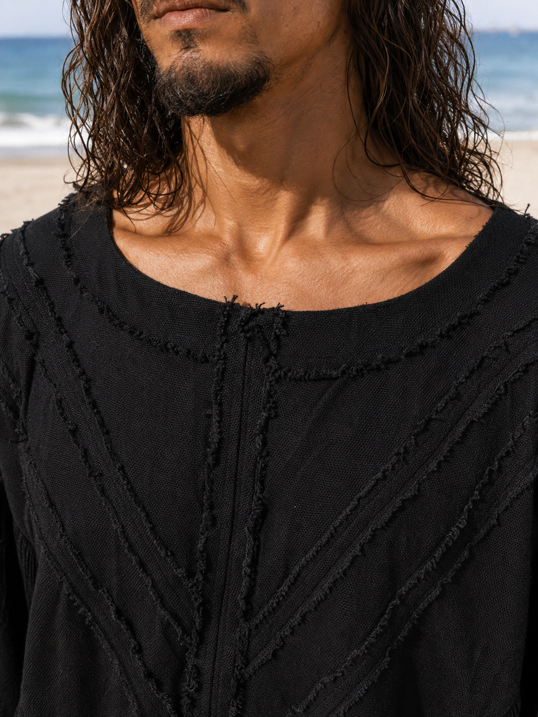 Close-up of a man wearing a black textured top with frayed trim, photographed outdoors on a beach with ocean and sand in the background.