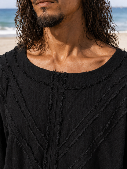 Close-up of a man wearing a black textured top with frayed trim, photographed outdoors on a beach with ocean and sand in the background.