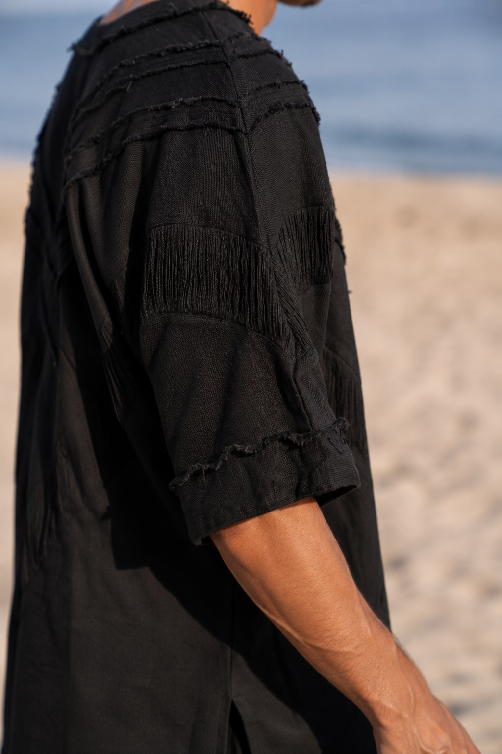A close-up side view of a person wearing a black textured short-sleeve top with frayed, layered details, standing on a sandy beach with the ocean in the background.