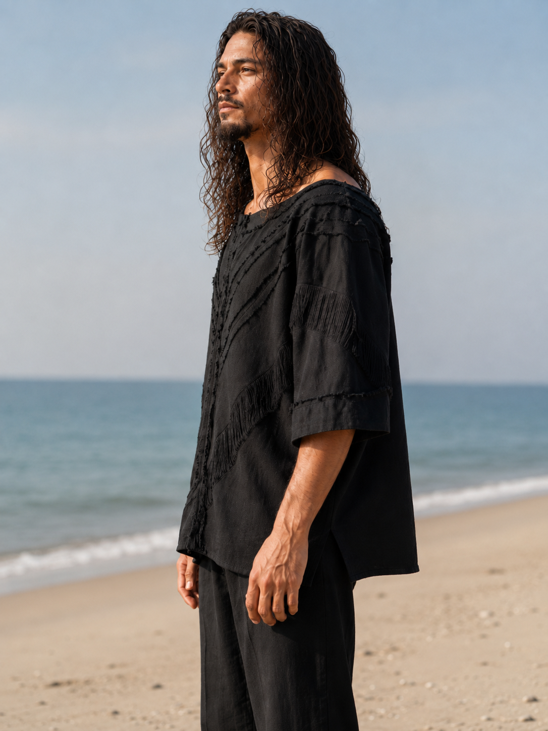 A man with long dark hair stands on a sandy beach wearing a loose black short-sleeve top and matching black pants, with the ocean and pale sky behind him.