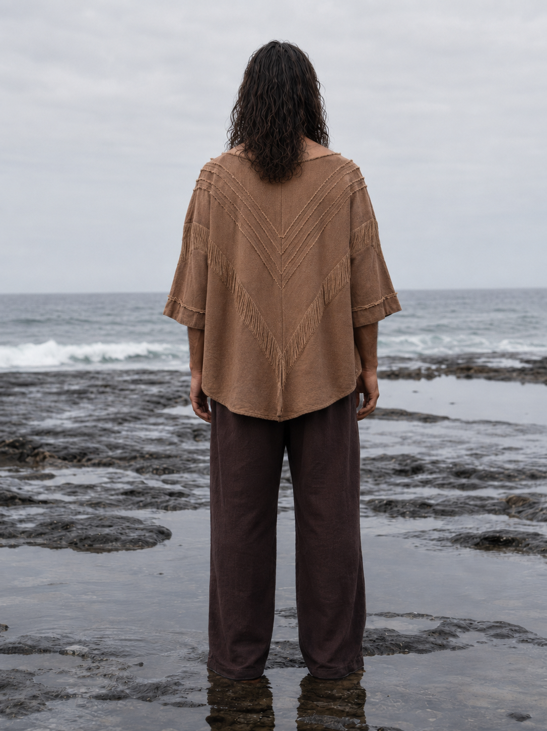 A person standing on a rocky ocean shore, viewed from behind, wearing a loose tan knit top with textured chevron detailing and dark brown wide-leg pants.