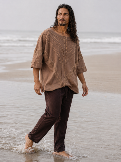 A man with long dark hair walks through shallow ocean water on a beach, wearing a loose brown embroidered tunic top and dark brown pants.