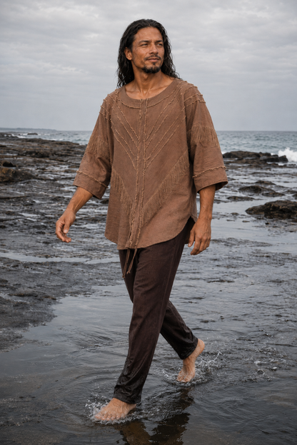 A man walking barefoot through shallow ocean water on a rocky beach, wearing a loose brown textured tunic top and dark brown pants.
