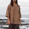 A man with long dark hair stands barefoot in shallow ocean water on a rocky shoreline, wearing a loose brown short-sleeve top and dark brown pants.