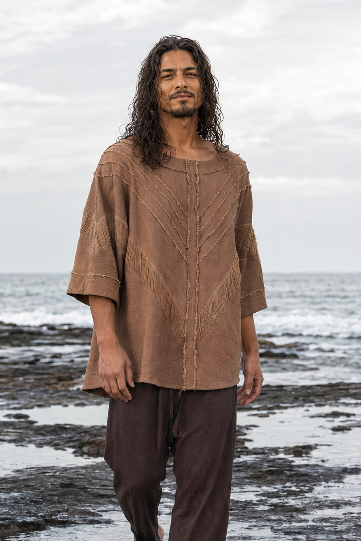 A man with long dark hair stands barefoot in shallow ocean water on a rocky shoreline, wearing a loose brown short-sleeve top and dark brown pants.